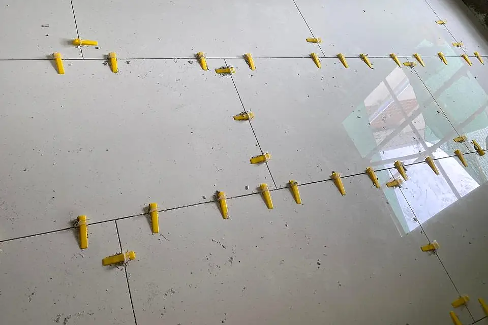 Glossy white floor tiles held level by rows of yellow levelling clips and wedges along every joint