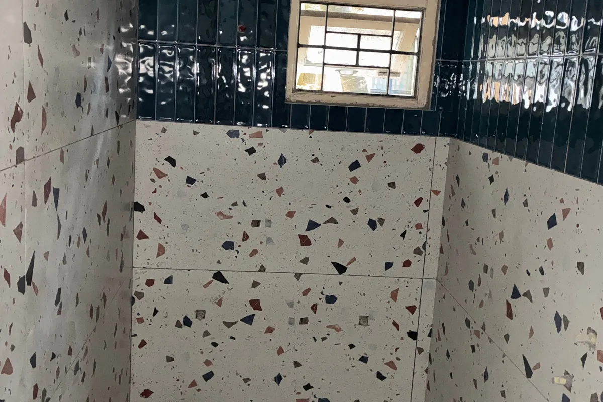 Bathroom walls and floor in white terrazzo-look tiles with coloured chips, under a band of glossy navy tiles around a small window