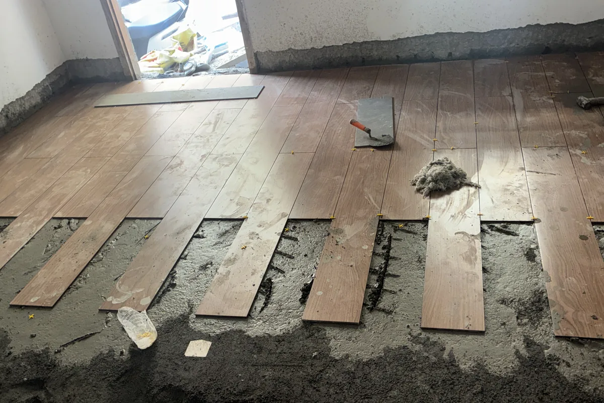 Wooden floor planks being laid on a mortar bed with a trowel in a room under renovation