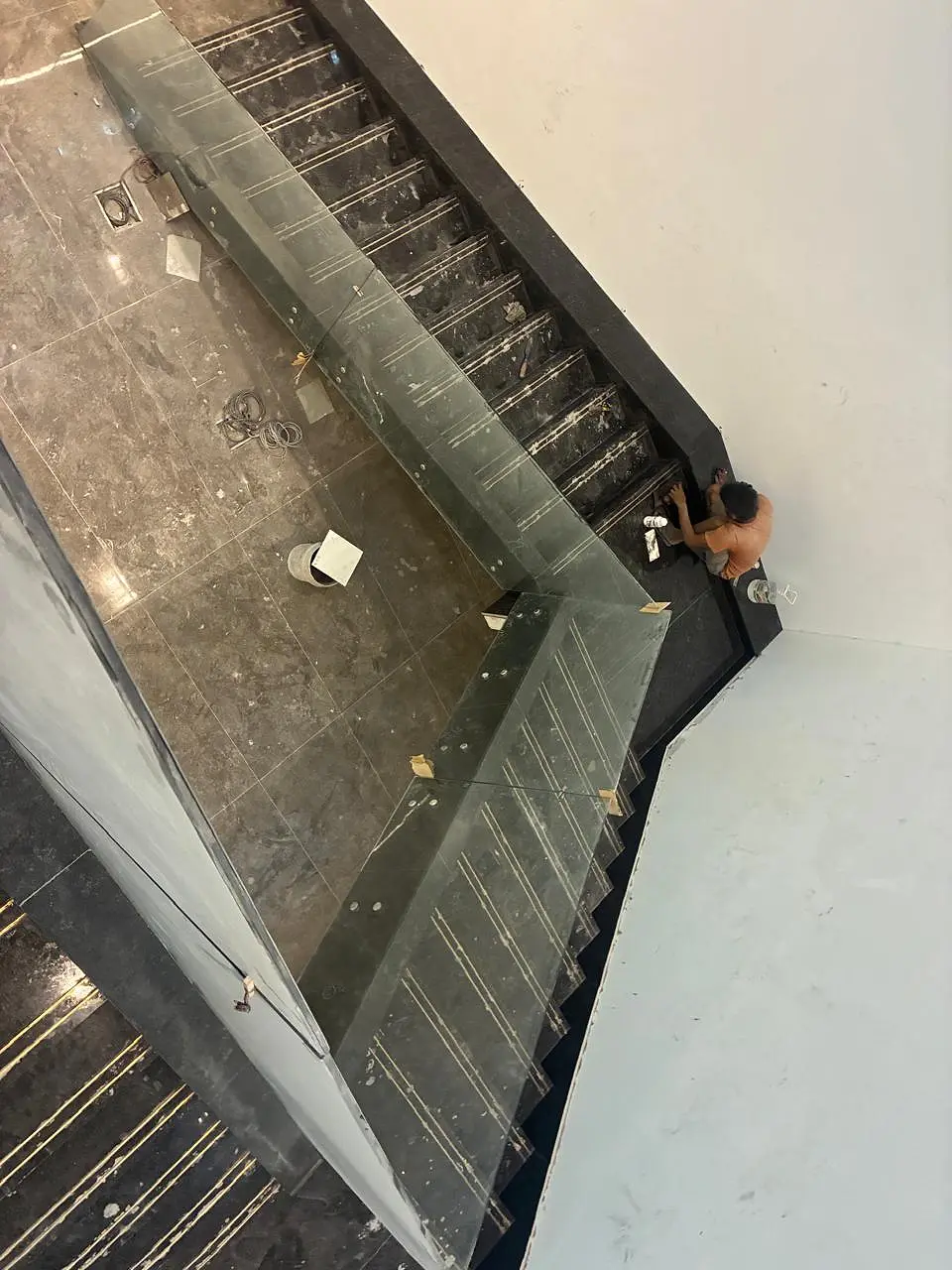 Overhead view down a stairwell: three runs of black granite stairs with brass strips wind down beside a frameless glass railing, a fitter working on a step